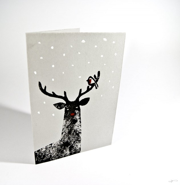 Photograph of a linocut reindeer printed in black, with a painted red nose, robin and white snow on grey paper, on a clean white background.
