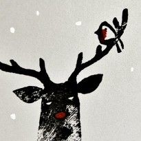 Photograph of a linocut reindeer printed in black, with a painted red nose, robin and white snow on grey paper, on a clean white background.